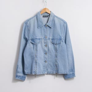 May include: Light blue denim jacket with a collared neckline, button-up front, and two chest pockets. The jacket features silver-toned stud detailing along the front and zippers at the hem. The jacket is hanging on a wooden hanger.