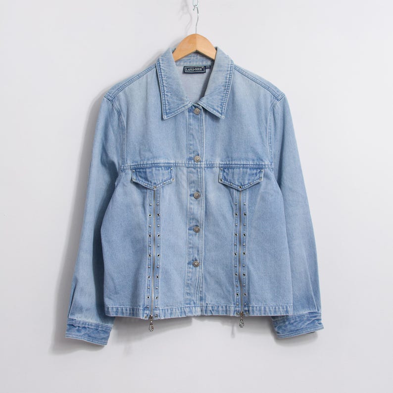 May include: Light blue denim jacket with a collared neckline, button-up front, and two chest pockets. The jacket features silver-toned stud detailing along the front and zippers at the hem. The jacket is hanging on a wooden hanger.