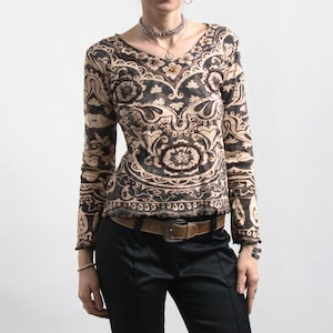 Könnte beinhalten: Ein langärmliges Oberteil mit V-Ausschnitt und einem braunen und beigefarbenen Paisley-Muster. Das Oberteil hat eine taillierte Silhouette und wird mit einer schwarzen Hose und einem braunen Gürtel kombiniert. Der Gesamtstil ist böhmisch.