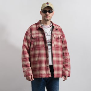 Pode incluir: Uma camisa de manga comprida abotoada com padrão xadrez vermelho e bege com dois bolsos no peito, usada sobre uma t-shirt branca com estampado gráfico, combinada com calças de ganga azuis, um boné bege com um pequeno logótipo de urso e óculos escuros.