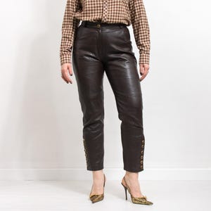 May include: Dark brown leather pants with gold button details along the ankles. The pants have a classic, tailored fit. The model is wearing a brown plaid button-down shirt and gold-colored heels.