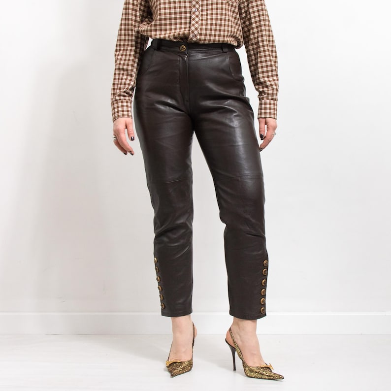 May include: Dark brown leather pants with gold button details along the ankles. The pants have a classic, tailored fit. The model is wearing a brown plaid button-down shirt and gold-colored heels.