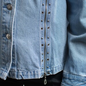May include: Light blue denim jacket with silver-tone metal studs and a full zipper closure. The jacket features two silver buttons on the left side and a decorative zipper pull. The jacket's design is a classic style.