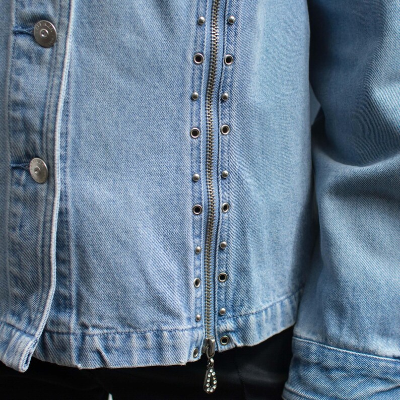 May include: Light blue denim jacket with silver-tone metal studs and a full zipper closure. The jacket features two silver buttons on the left side and a decorative zipper pull. The jacket's design is a classic style.