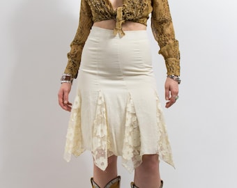 May include: Cream-colored skirt with lace detailing at the hem. The skirt has a high waist and a flared design. The model is wearing a brown patterned top and cowboy boots. The skirt is a vintage style.