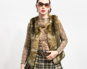 May include: A woman wearing a brown faux fur vest over a brown and beige patterned mesh top. She is also wearing a brown leather belt with a silver buckle, a plaid skirt, and sunglasses.