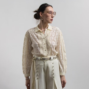 Peut inclure: Chemisier en dentelle crème à manches longues et fermeture boutonnée. Il est associé à un pantalon blanc cassé et une ceinture décorative. L'ensemble est complété par des lunettes et des bijoux, créant une esthétique vintage.