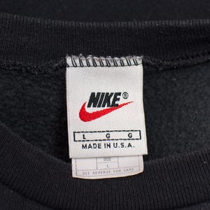 May include: Close-up of a black sweatshirt collar with a white tag. The tag features the Nike logo in red and white, with the text "NIKE" and "MADE IN U.S.A.". The size is L.
