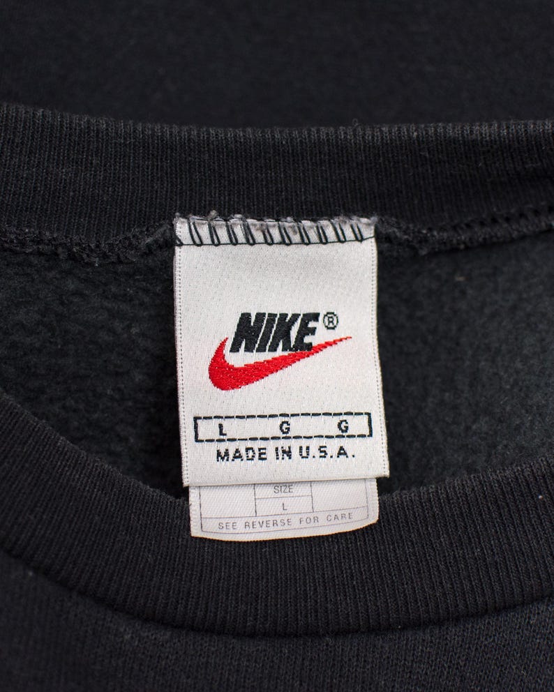 May include: Close-up of a black sweatshirt collar with a white tag. The tag features the Nike logo in red and white, with the text "NIKE" and "MADE IN U.S.A.". The size is L.