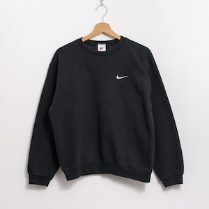 May include: Black crew neck sweatshirt with a small white Nike swoosh logo on the left chest and a small Nike tag on the upper chest. The sweatshirt is hanging on a wooden hanger.