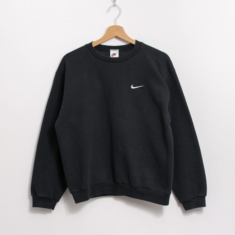 May include: Black crew neck sweatshirt with a small white Nike swoosh logo on the left chest and a small Nike tag on the upper chest. The sweatshirt is hanging on a wooden hanger.