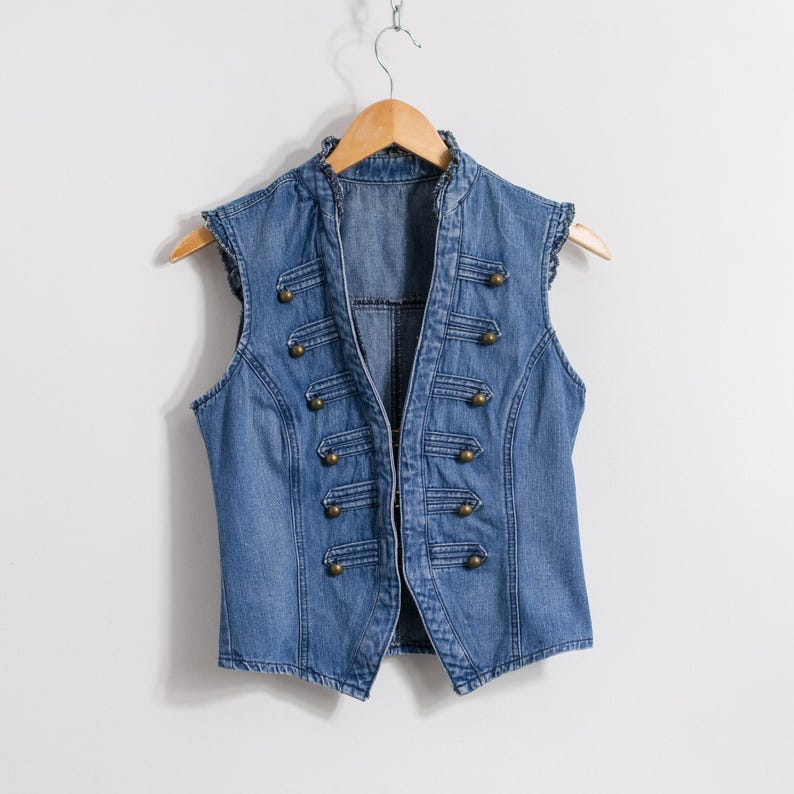 May include: A sleeveless blue denim vest featuring a high collar and a military-inspired front design. The garment includes five pairs of decorative brass-toned buttons arranged vertically on each side, connected by horizontal stitched cord detailing.