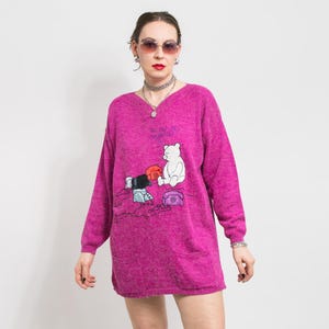 May include: A pink long-sleeved sweater with a cartoon bear and phone design. The sweater has a V-neck and a pocket on the front. The text on the sweater reads "We'll get you connected".