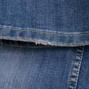 Peut inclure: Gros plan sur un tissu denim bleu avec un bord effiloch&eacute;. L'image montre la texture du denim, les coutures et une petite zone effiloch&eacute;e. Le denim est de couleur bleu moyen.