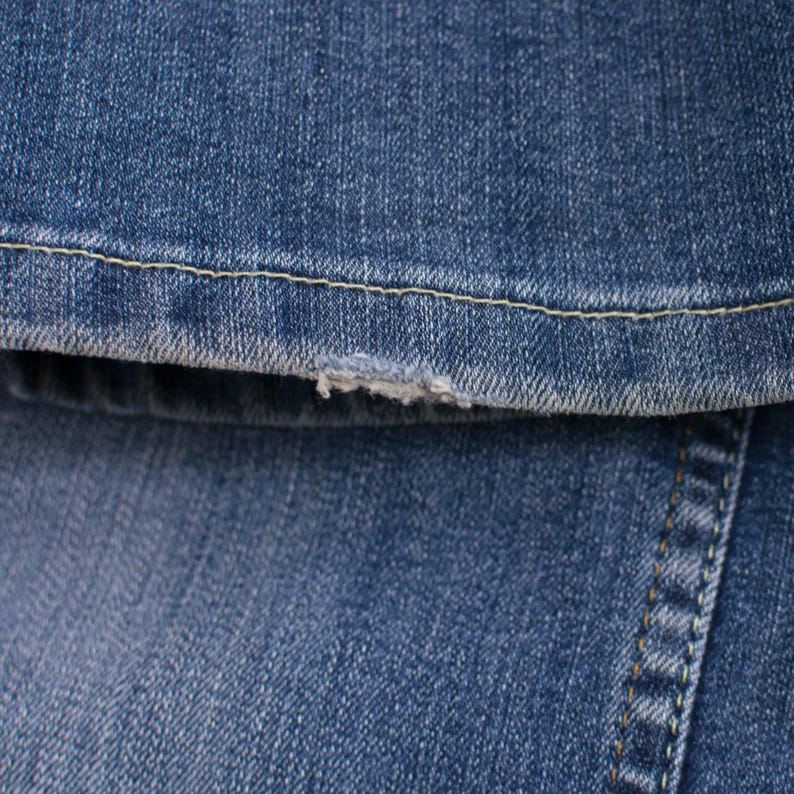 Peut inclure: Gros plan sur un tissu denim bleu avec un bord effiloch&eacute;. L'image montre la texture du denim, les coutures et une petite zone effiloch&eacute;e. Le denim est de couleur bleu moyen.