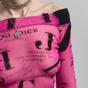 Pu&ograve; includere: Un top rosa acceso, con spalle scoperte, con dettagli neri e un rivestimento in rete. Il top presenta un motivo ripetuto di testo nero e uno scollo a coste nero. &Egrave; visibile una collana d'argento.