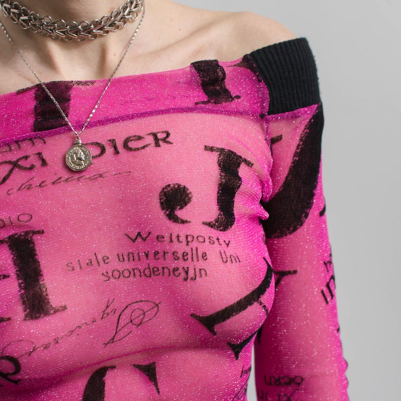 Pu&ograve; includere: Un top rosa acceso, con spalle scoperte, con dettagli neri e un rivestimento in rete. Il top presenta un motivo ripetuto di testo nero e uno scollo a coste nero. &Egrave; visibile una collana d'argento.