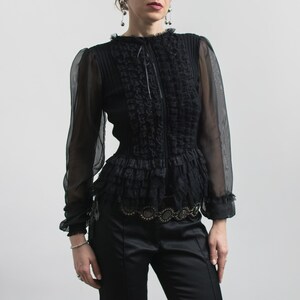 Puede incluir: Blusa negra con mangas largas transparentes, cuello con volantes y cintura entallada. El corpi&ntilde;o presenta detalles texturizados intrincados y un lazo delicado en el escote. La blusa se combina con pantalones de sat&eacute;n negros.