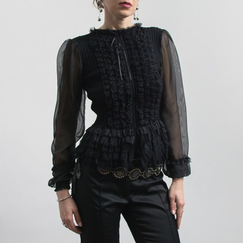 Puede incluir: Blusa negra con mangas largas transparentes, cuello con volantes y cintura entallada. El corpi&ntilde;o presenta detalles texturizados intrincados y un lazo delicado en el escote. La blusa se combina con pantalones de sat&eacute;n negros.