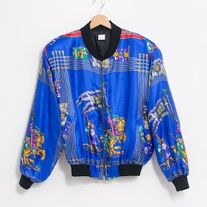 May include: A blue satin bomber jacket with a zipper closure and a colorful patterned design featuring knights on horseback. The jacket has a black ribbed collar, cuffs, and waistband.