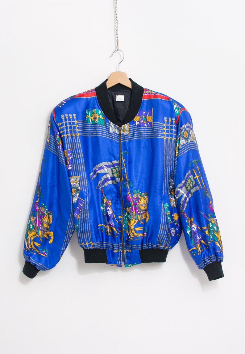 May include: A blue satin bomber jacket with a zipper closure and a colorful patterned design featuring knights on horseback. The jacket has a black ribbed collar, cuffs, and waistband.