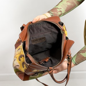May include: A brown leather handbag with a colorful floral pattern on the side. The bag has a zipper closure and a brown leather strap. The interior is black with a small pocket and the brand name "Russell Bromley" is visible.