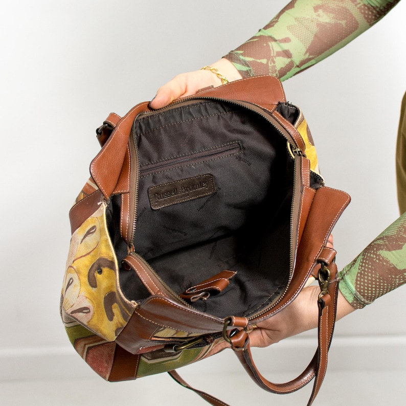 May include: A brown leather handbag with a colorful floral pattern on the side. The bag has a zipper closure and a brown leather strap. The interior is black with a small pocket and the brand name "Russell Bromley" is visible.