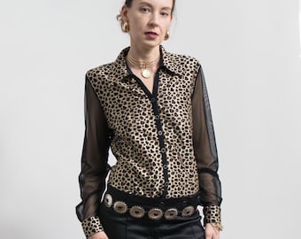 Op de afbeelding: Een button-down shirt met een luipaardprintpatroon. De blouse heeft zwarte mesh mouwen en een zwarte kraag. Het model draagt een zwarte riem met zilveren versieringen en een zwarte broek. Het model draagt ook gouden sieraden.