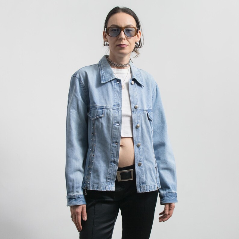 May include: Light blue denim jacket with silver stud accents, worn over a white crop top and black pants. The jacket has a classic collar, button closures, and two chest pockets. The person is wearing sunglasses and a silver necklace.