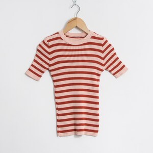 May include: A short-sleeved, ribbed knit top with horizontal stripes in light pink and reddish-brown. The top is suspended from a wooden hanger against a white backdrop. The neckline and sleeve cuffs are light pink.