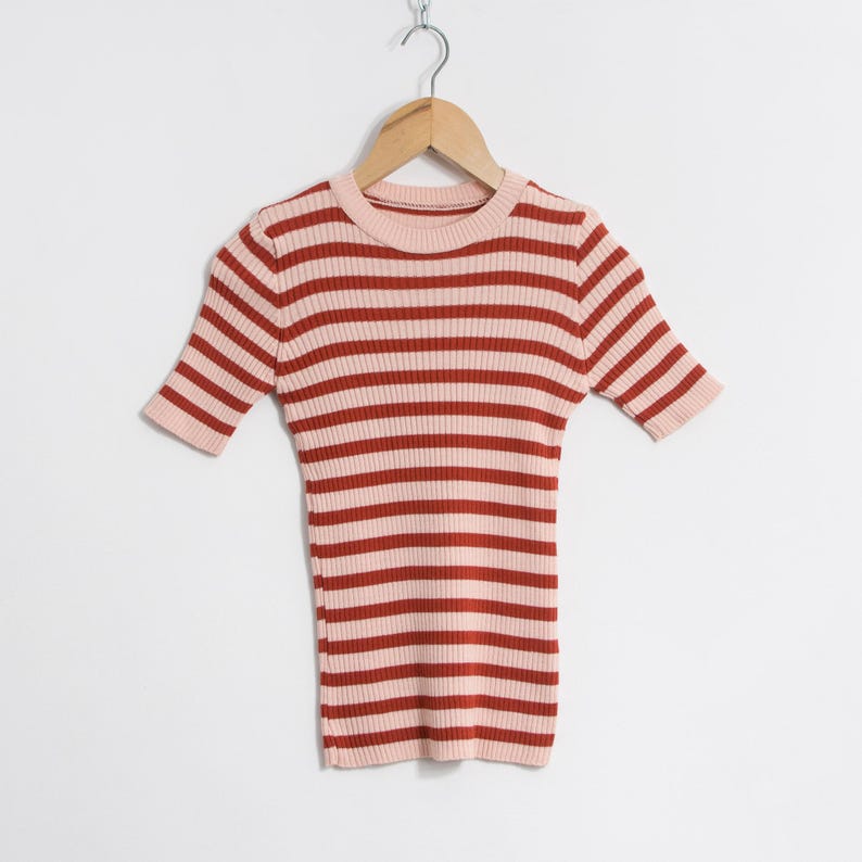 May include: A short-sleeved, ribbed knit top with horizontal stripes in light pink and reddish-brown. The top is suspended from a wooden hanger against a white backdrop. The neckline and sleeve cuffs are light pink.