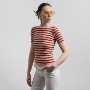 May include: A short-sleeved, horizontally striped knit top in shades of red and pink. The top is paired with white trousers and a silver chain belt. The person is wearing orange-tinted sunglasses and silver jewellery.