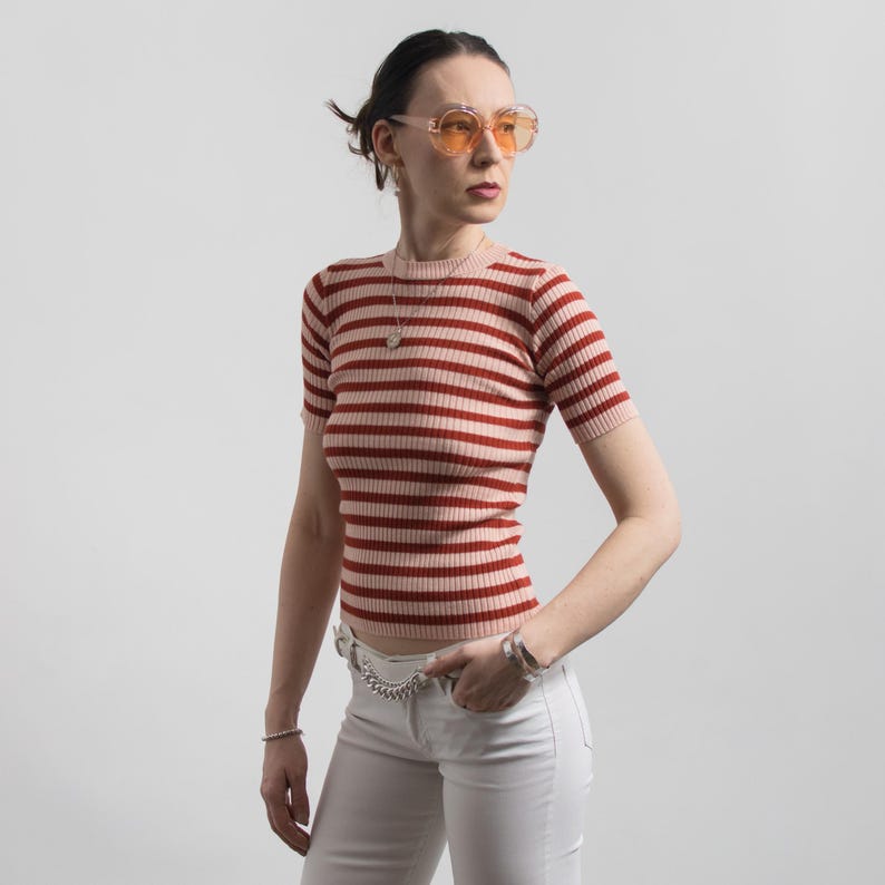 May include: A short-sleeved, horizontally striped knit top in shades of red and pink. The top is paired with white trousers and a silver chain belt. The person is wearing orange-tinted sunglasses and silver jewellery.