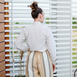 May include: A woman wearing a white button-down shirt and striped pants stands in front of a window with white blinds.