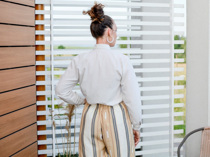 May include: A woman wearing a white button-down shirt and striped pants stands in front of a window with white blinds.