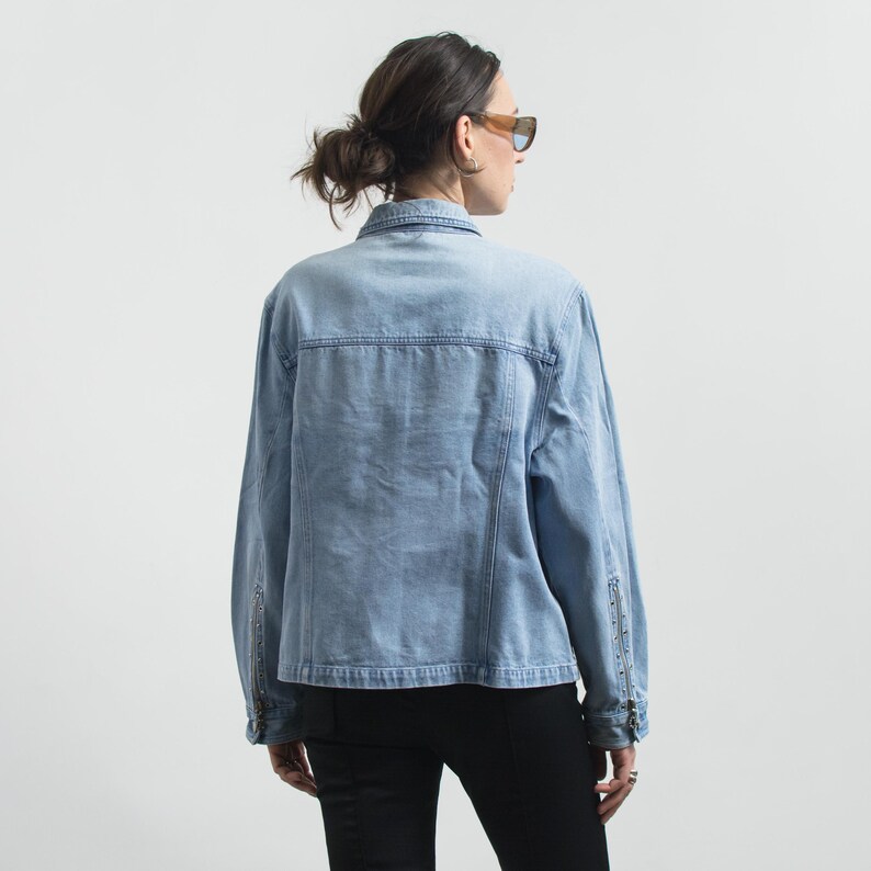 May include: A light blue denim jacket featuring a classic collar, long sleeves with vertical zippered cuffs, and silver-toned metal stud detailing along the sleeve zippers. The jacket has a structured back yoke and visible seam stitching.