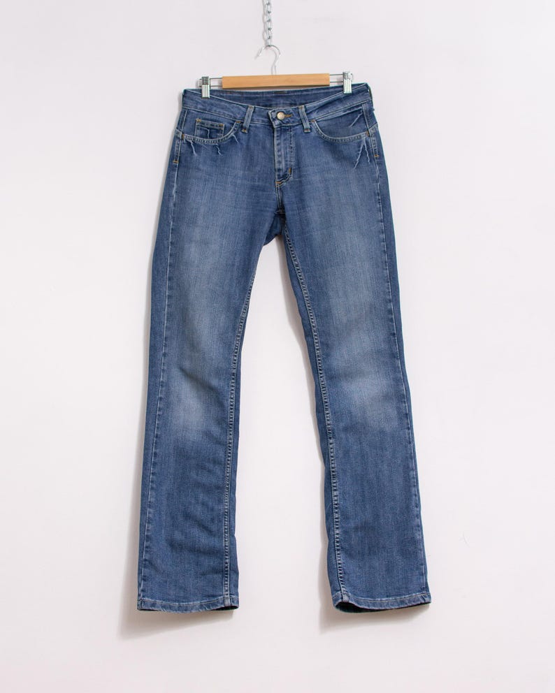 Peut inclure: Un jean en denim bleu suspendu &agrave; un cintre en bois. Le jean a une coupe droite classique &agrave; cinq poches, une fermeture &agrave; bouton et fermeture &eacute;clair. Le denim pr&eacute;sente un d&eacute;lavage d&eacute;color&eacute; avec des d&eacute;tails subtils.