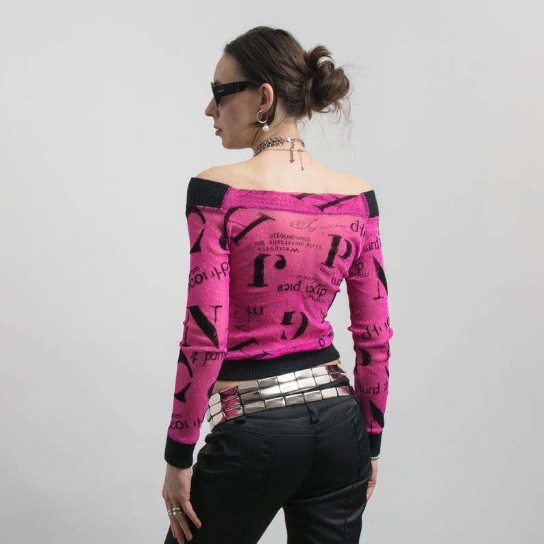 Pu&ograve; includere: Un top rosa acceso con spalle scoperte, rifiniture nere e maniche lunghe. Il top presenta un motivo di testo nero ripetuto. La modella indossa pantaloni neri e una cintura argentata. Lo stile generale &egrave; alla moda.