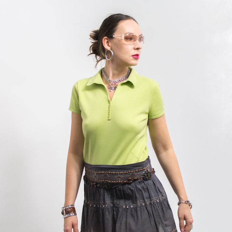 K&ouml;nnte beinhalten: Eine Frau tr&auml;gt ein hellgr&uuml;nes Poloshirt mit einem braunen Perleng&uuml;rtel und einen grauen Rock. Sie tr&auml;gt eine klare Brille und silbernen Schmuck.