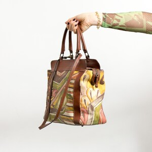 May include: A brown leather handbag with a colorful abstract design. The bag features a top handle, a shoulder strap, and a zipper closure. The design includes shades of yellow, green, brown, and white.