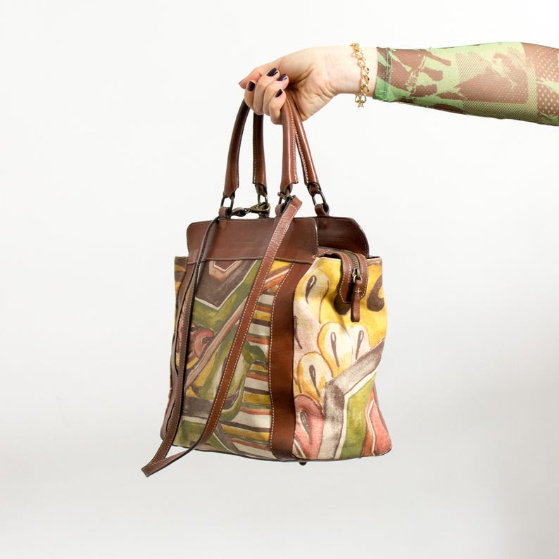 May include: A brown leather handbag with a colorful abstract design. The bag features a top handle, a shoulder strap, and a zipper closure. The design includes shades of yellow, green, brown, and white.