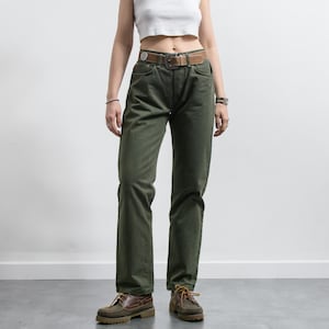 Op de afbeelding: Olijfgroene spijkerbroek met een bruine leren riem. De jeans heeft een klassiek ontwerp met vijf zakken en een rechte pijp. De persoon draagt bruine leren schoenen met veters. De outfit is eenvoudig en casual.