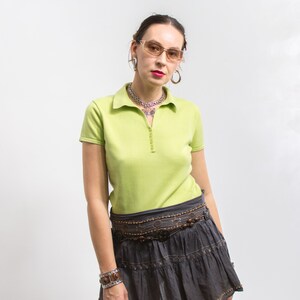 K&ouml;nnte beinhalten: Eine Frau tr&auml;gt ein limegr&uuml;nes Poloshirt mit einer silbernen Kettenkette und einem braunen Perleng&uuml;rtel. Sie tr&auml;gt einen grauen Rock mit einem geschichteten Design und braunen Perlenakzenten. Sie tr&auml;gt auch eine braune Brille mit Rahmen und Creolen.
