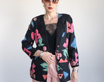 May include: Black blazer with a colorful floral pattern in pink, blue, and green. The blazer has a classic lapel and button closure. The model is wearing a black top and pink pants.