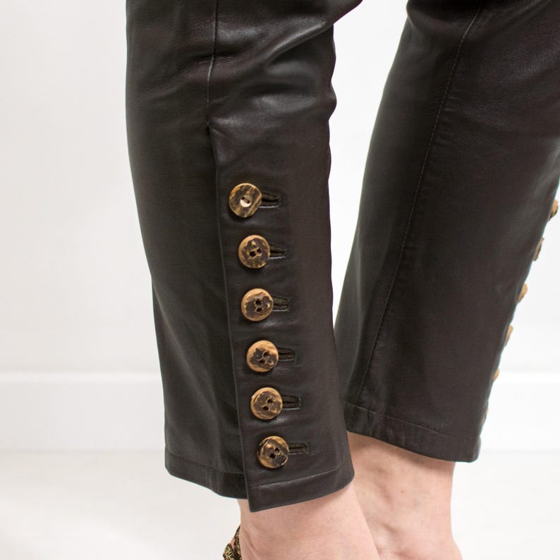 May include: Cropped brown leather pants with decorative wooden buttons running up the outer seam. The pants are a dark brown color and appear to be made of soft leather. The buttons are round and have a natural wood grain.