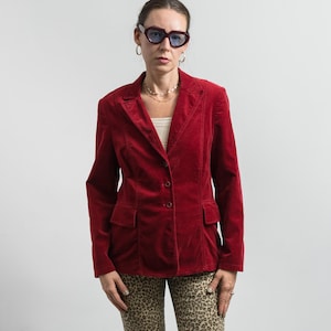 Puede incluir: Una americana de pana de color rojo intenso con solapa de muesca, tres botones frontales y dos bolsillos con solapa. La americana se lleva sobre una prenda superior blanca y se combina con pantalones de estampado de leopardo beige y negro.