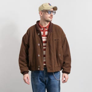 May include: A brown bomber jacket with a button-up front, side pockets, and a brown collar. The jacket is worn over a red and white striped polo shirt and blue jeans. A baseball cap and sunglasses complete the look.
