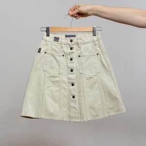 May include: A light beige denim skirt with a button-up front and two front pockets. The skirt is held up by a hanger. The brand name "VERSACE" is visible on the waistband. The skirt has a classic, A-line silhouette.