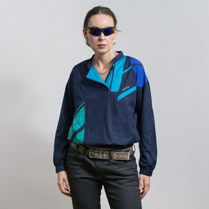 Puede incluir: Chaqueta Adidas azul marino con detalles geométricos turquesa y azules. La chaqueta tiene cierre de cremallera de un cuarto y cuello alzado. La modelo lleva vaqueros negros y gafas de sol.