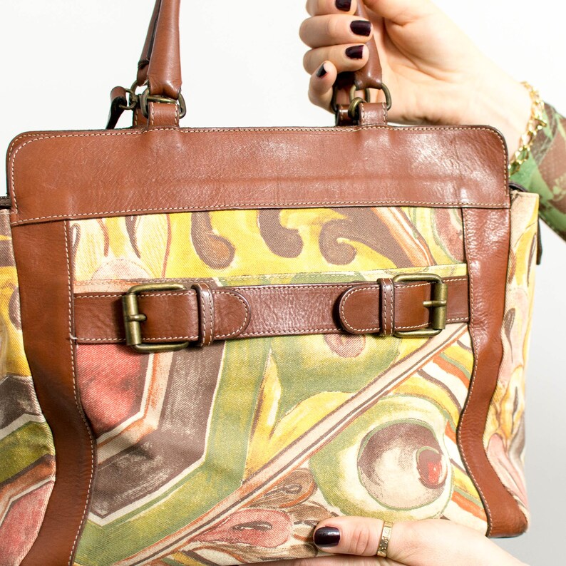 May include: A brown leather handbag with a colorful abstract pattern. The bag features brown leather trim, handles, and straps with brass buckles. The main body of the bag has a vibrant print with yellow, green, and red hues.